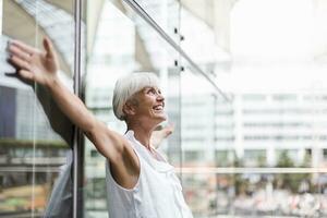 Happy senior woman leaning against glass facade with outstretched arms photo